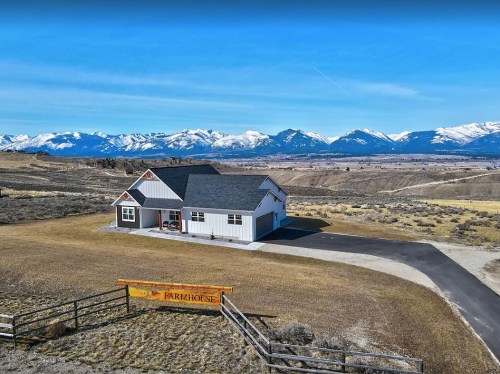 A modern house with a gray roof sits on a hillside, surrounded by mountains and open fields under a clear blue sky.