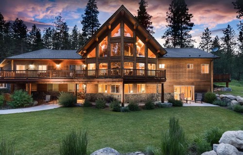 A large wooden house with large windows, surrounded by trees and a grassy area, under a colorful sunset sky.