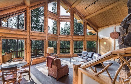 Cozy cabin interior with large windows, wooden beams, and a stone fireplace, surrounded by trees.