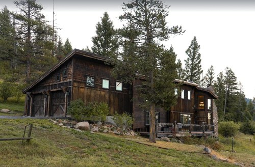 A rustic wooden house surrounded by trees and greenery, set in a mountainous landscape.