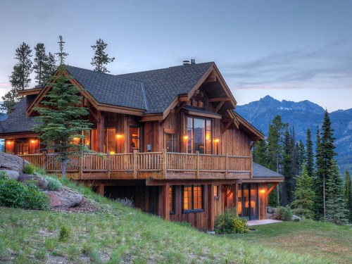 A wooden cabin with large windows, surrounded by trees and mountains, illuminated at dusk.