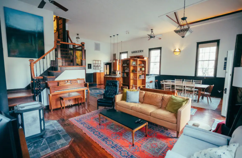 Spacious living area with a piano, cozy seating, and a dining table, featuring wooden floors and bright natural light.