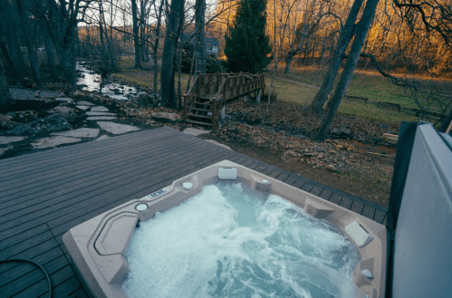 A hot tub on a wooden deck, surrounded by trees and a stream, with a bridge in the background.