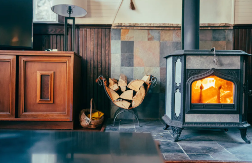 Cozy living room with a wood stove, stacked firewood, and a lamp, creating a warm atmosphere.
