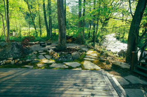 A serene outdoor scene with a wooden deck, stone path, and lush greenery surrounding a small fire pit area.