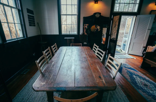 A rustic dining room with a large wooden table and chairs, featuring windows and an open door to the outdoors.