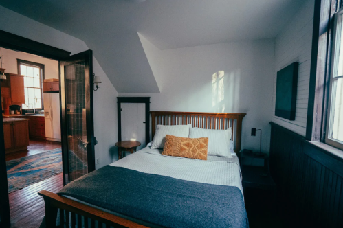 Cozy bedroom with a wooden bed, a side table, and sunlight streaming through the windows. Kitchen visible in the background.