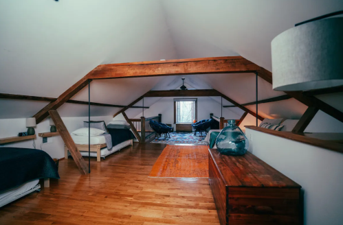 Spacious attic room with wooden beams, two beds, a rug, and seating area by a window. Warm, inviting atmosphere.