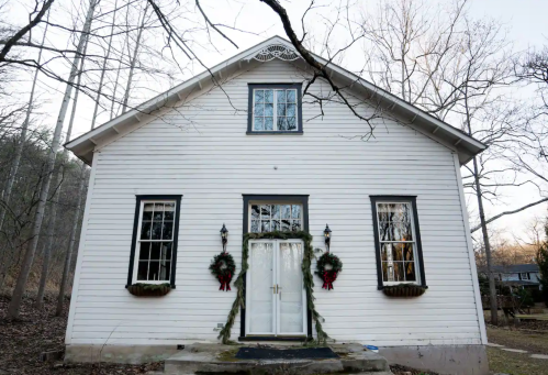 A white house with a gabled roof, featuring two wreaths on the front doors and surrounded by bare trees.