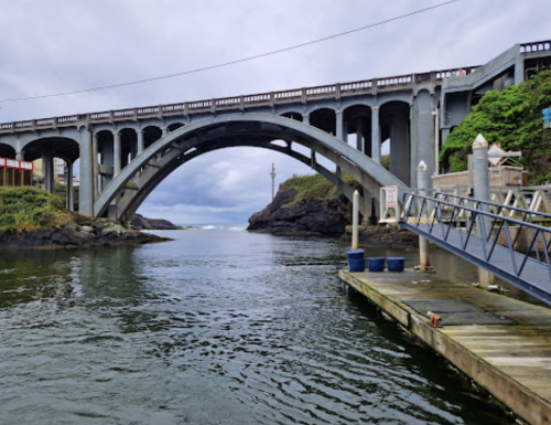 A concrete arch bridge spans a calm waterway, with a wooden dock and rocky shoreline in the foreground.
