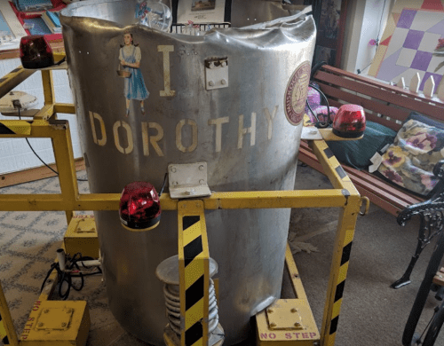 A large metallic structure with "I DOROTHY" written on it, surrounded by red lights and yellow caution barriers.