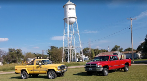 Two pickup trucks, one yellow and one red, parked on grass with a water tower in the background under a blue sky.