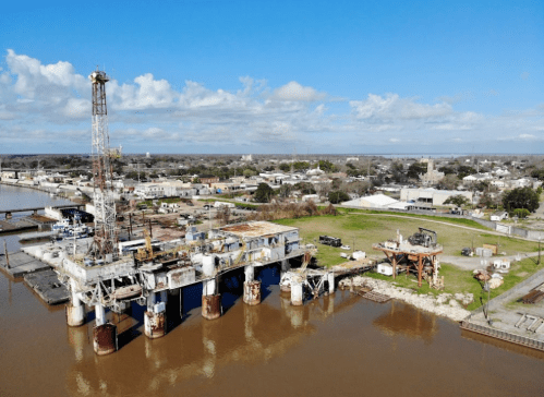 Aerial view of an oil rig near a waterfront, surrounded by industrial buildings and a clear blue sky.