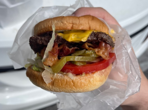 A hand holding a burger with a beef patty, cheese, bacon, lettuce, tomato, and green peppers, wrapped in paper.