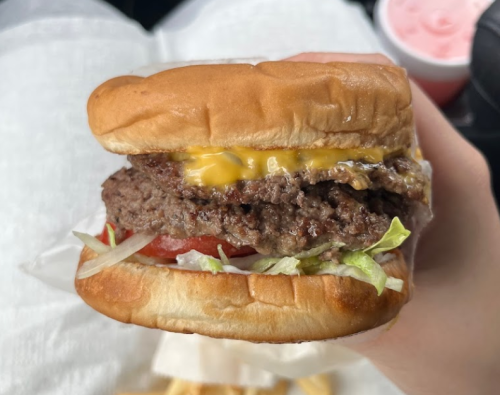A hand holding a cheeseburger with lettuce, tomato, and a juicy beef patty, with fries and a drink in the background.
