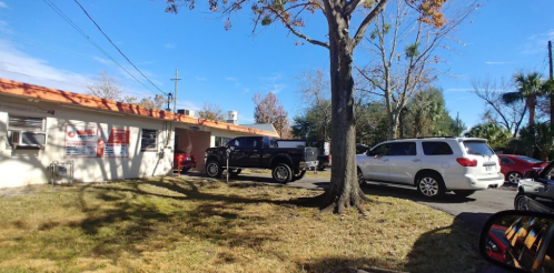 A parking area with a black truck and a white SUV near a building, surrounded by trees and clear blue skies.
