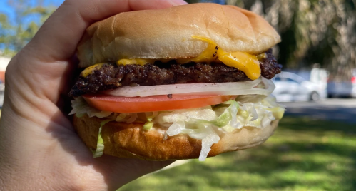 A hand holding a cheeseburger with lettuce, tomato, and onion against a blurred outdoor background.