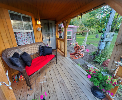 A cozy porch with a wicker sofa, red cushions, and a hammock, surrounded by greenery and colorful flowers.