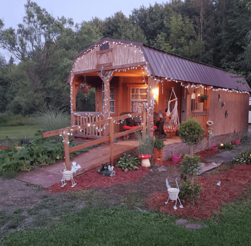 A cozy wooden cabin adorned with string lights, surrounded by greenery and colorful flowers in a serene outdoor setting.