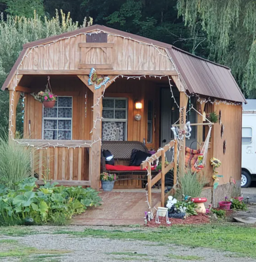 A cozy wooden cabin with a porch, decorated with plants and lights, surrounded by greenery.