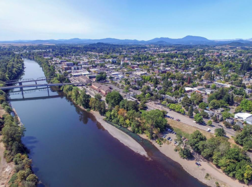 Aerial view of a river winding through a town, surrounded by trees and mountains in the background under a clear blue sky.