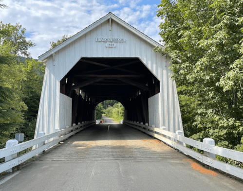 A white covered bridge with a sign reading "Hayden Bridge 1918," surrounded by greenery and a clear blue sky.