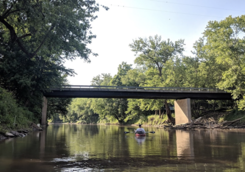 A calm river scene with a bridge overhead and a small boat gliding through the water, surrounded by lush greenery.