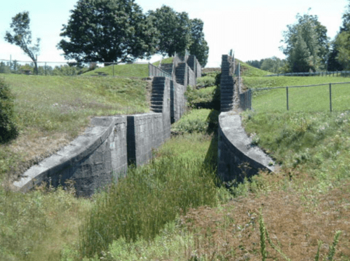 A stone structure with curved walls, surrounded by grass and trees, set in a sunny landscape.