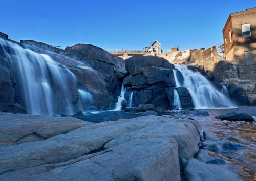 A serene waterfall cascades over large rocks, with a clear blue sky and a building in the background.
