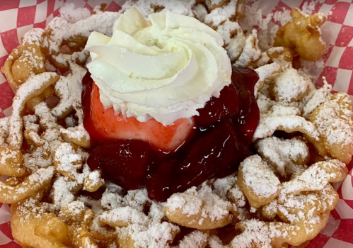 A funnel cake topped with whipped cream, strawberry sauce, and a cherry, dusted with powdered sugar.