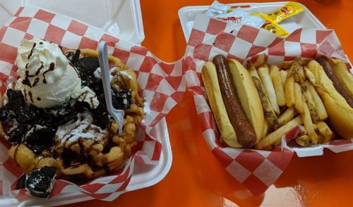 A plate of funnel cake topped with whipped cream and chocolate, alongside two hot dogs and fries in a takeout container.