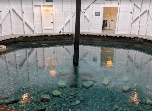 A circular pool with clear water and visible stones, surrounded by white wooden walls and a doorway in the background.