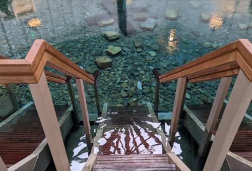 Wooden stairs lead down to a serene pool with clear water and smooth stones at the bottom.
