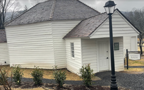 A small white building with a sloped roof, labeled "Gentlemen's Bath," beside a lamp post and landscaped area.