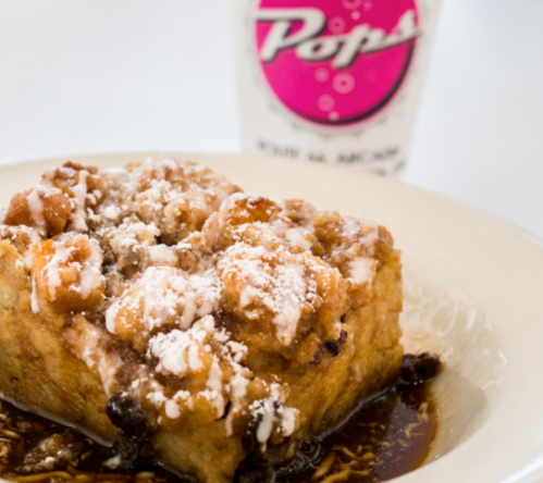 A slice of dessert topped with powdered sugar and chocolate sauce, served on a plate with a drink in the background.