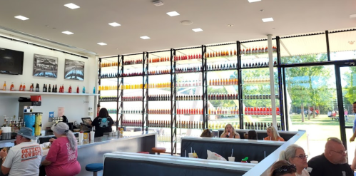 Bright, modern restaurant interior with colorful condiment shelves, patrons seated at tables, and large windows.