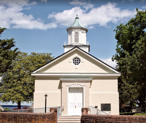 A small, white church with a green roof and steeple, surrounded by trees under a partly cloudy sky.