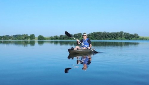 A person paddles a kayak on a calm lake, surrounded by greenery under a clear blue sky.