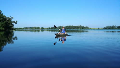 A person paddles a kayak on a calm lake, surrounded by trees and clear blue skies.