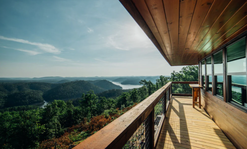A wooden balcony overlooks a scenic view of mountains and a lake under a clear blue sky.