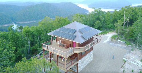 A modern, eco-friendly house with solar panels, surrounded by lush greenery and overlooking a lake and mountains.