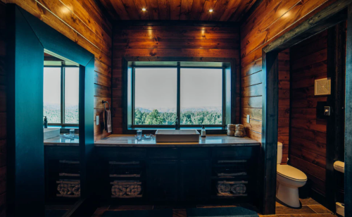 A rustic bathroom with wooden walls, a large window, a sink, and a toilet, offering a scenic view of nature outside.