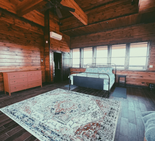 Cozy wooden bedroom with a bed, dresser, and a large patterned rug, featuring large windows for natural light.