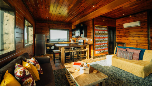 Cozy wooden cabin interior with a living area, kitchen, and large windows showcasing scenic views. Warm, inviting decor.