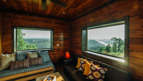 Cozy wooden cabin interior with large windows showcasing a scenic view of mountains and a lake.
