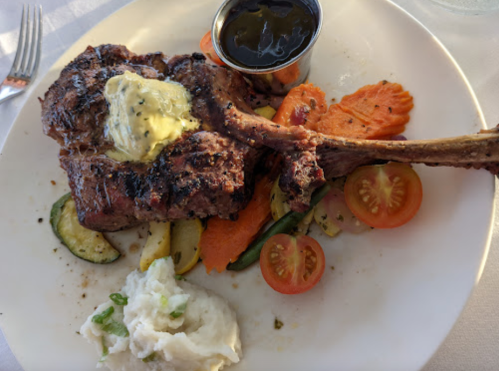 A plate with a grilled steak topped with butter, served with vegetables, mashed potatoes, and a small cup of sauce.