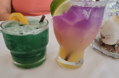Two colorful cocktails on a table: one green with a lemon twist, the other purple and orange with a lime wedge.