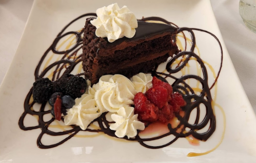 A slice of chocolate cake topped with whipped cream, surrounded by berries and drizzled with chocolate sauce.