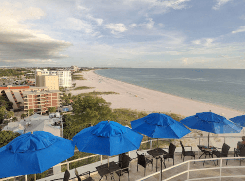 A scenic beach view with blue umbrellas and chairs overlooking a sandy shoreline and calm ocean under a cloudy sky.