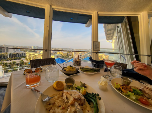 A dining table with a view of the city, featuring plates of food, drinks, and a scenic backdrop through large windows.